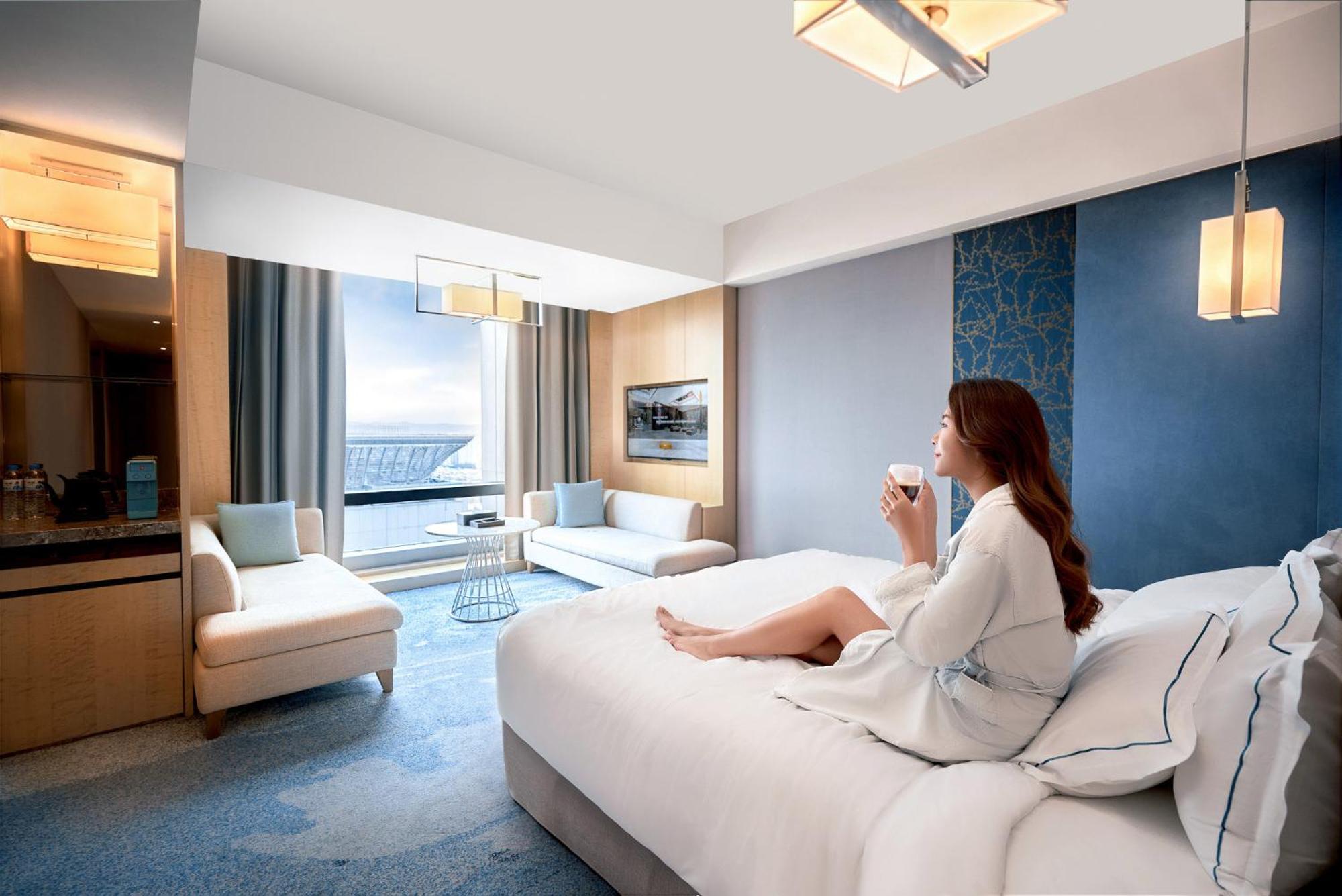 臺北 板桥凯撒大饭店酒店 外观 照片 The photo depicts a modern hotel room featuring a large bed with a light-colored blanket and pillows. A woman in a white robe is sitting on the bed, holding a cup, and appears to be enjoying the serene environment. The room has large windows that off