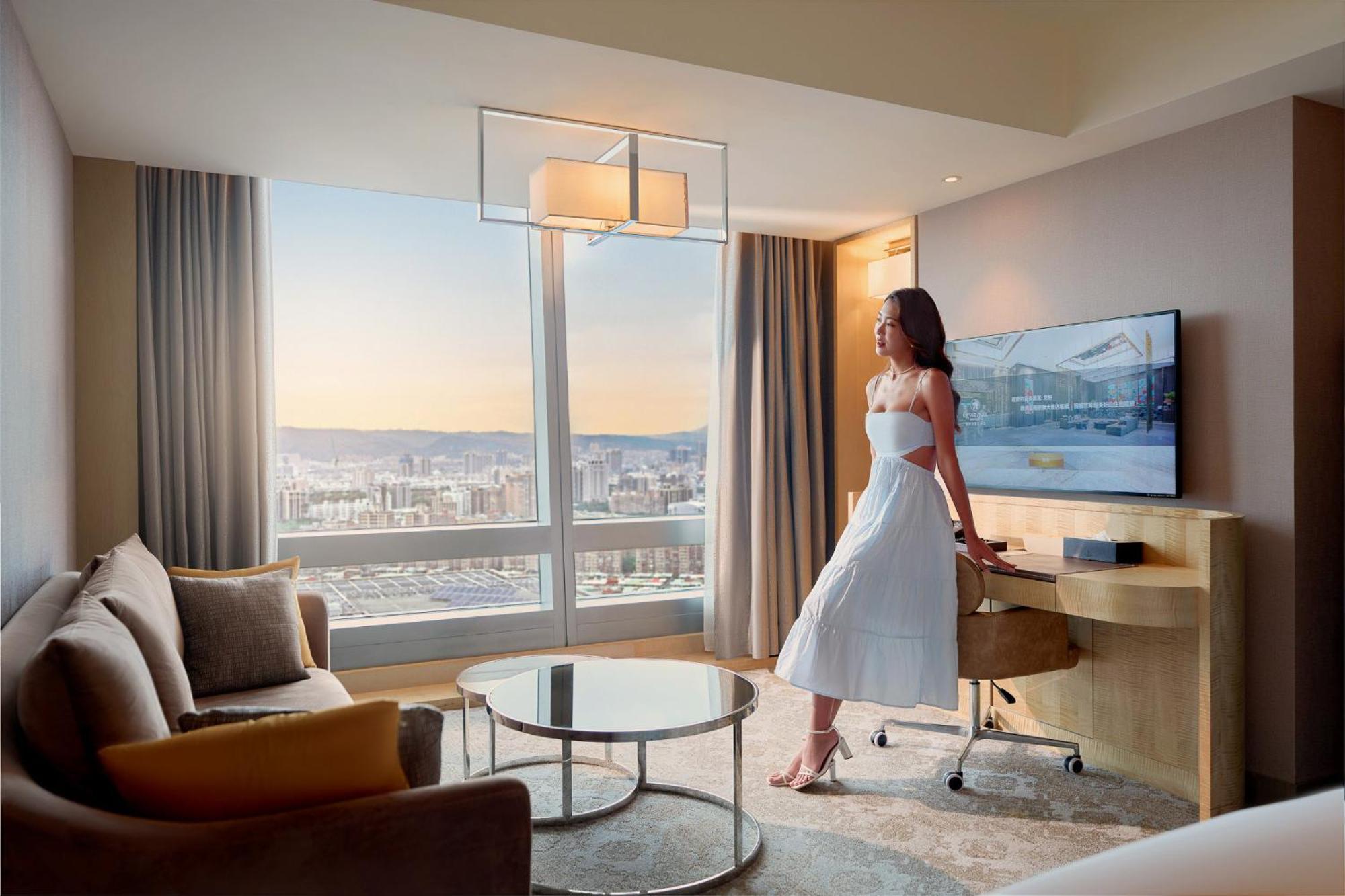 臺北 板桥凯撒大饭店酒店 外观 照片 The photo showcases a modern hotel room with a panoramic view of the city. A woman in a white dress stands next to a stylish table, gazing out of a large window. The room features contemporary furniture, including a sofa, a glass coffee table, and a 