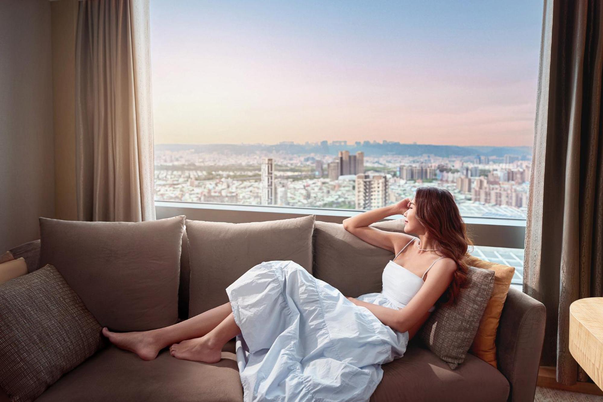 臺北 板桥凯撒大饭店酒店 外观 照片 The image shows a woman sitting on a couch in a bright room, gazing out of a large window that offers a view of a cityscape. She is wearing a white dress and appears to be in a relaxed pose, resting her arm on her head. The window shows a pastel sky,