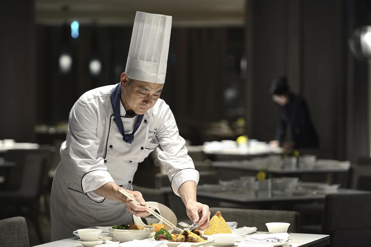 臺北 板桥凯撒大饭店酒店 外观 照片 The photo shows a chef in a professional kitchen setting. The chef, wearing a traditional white jacket and a tall chef's hat, is focused on plating a dish. There are various food items on the table, suggesting an elaborate meal is being prepared. In 