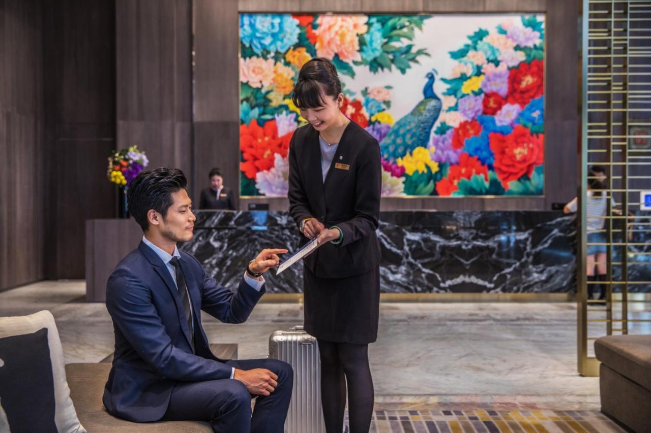 臺北 板桥凯撒大饭店酒店 外观 照片 The photo depicts a hotel lobby scene. In the foreground, a man in a tailored suit sits on a modern sofa, while a female staff member in a professional outfit is standing next to him, pointing at a tablet or document. The background features a vibran