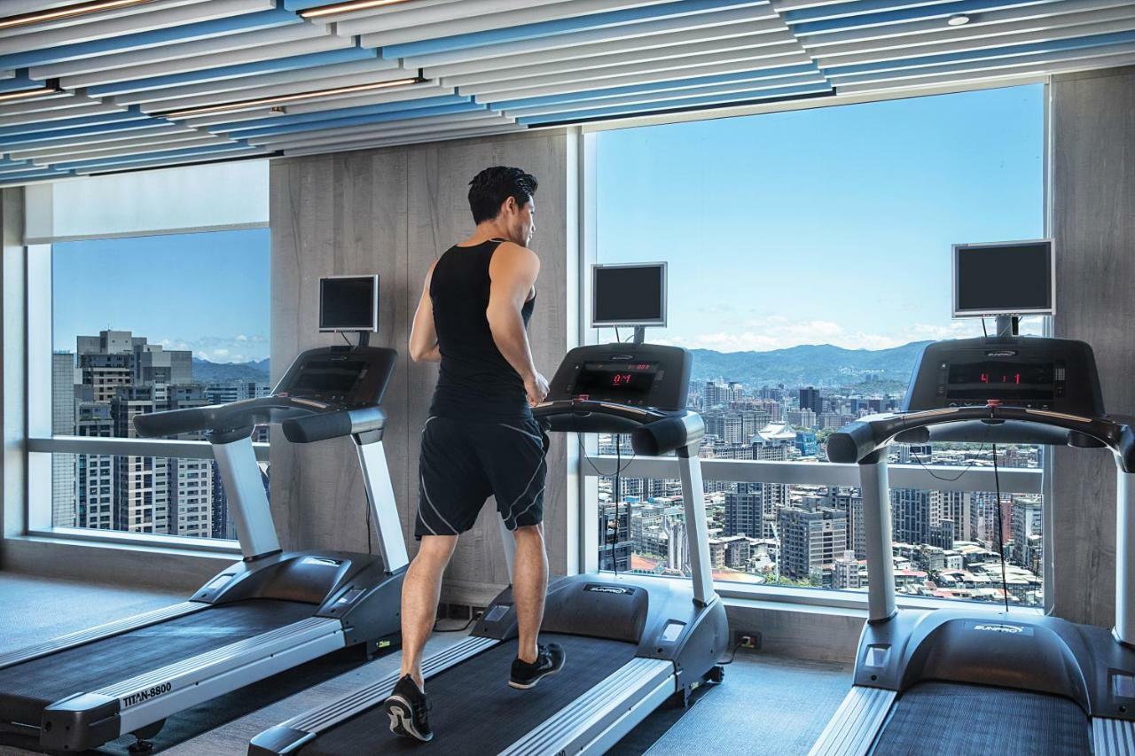 臺北 板桥凯撒大饭店酒店 外观 照片 The photo depicts a man running on a treadmill in a modern gym. He is wearing a sleeveless shirt and shorts, and appears to be focused on his workout. The gym has large windows that offer a panoramic view of a city skyline with distant mountains. The